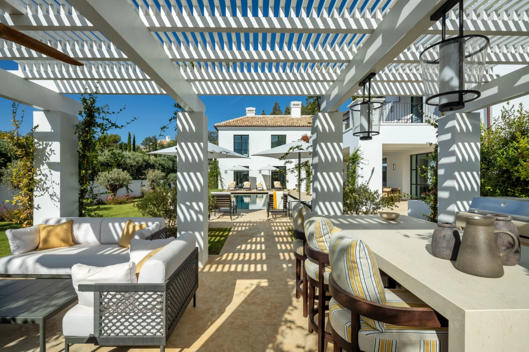 Shaded pergola terrace with outdoor dining, looking across the pool to a white Andalusian villa